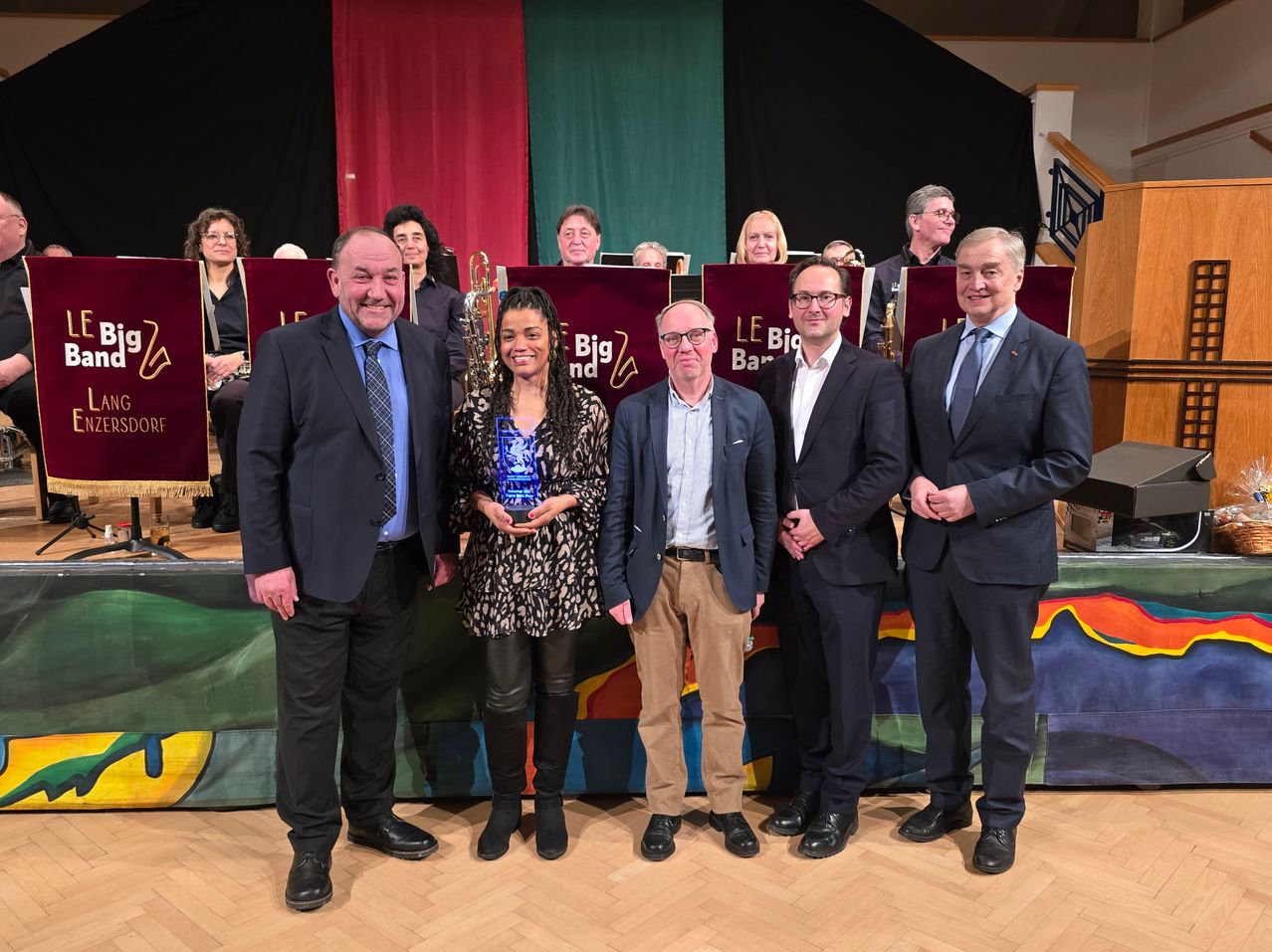 Eine Gruppe von Menschen steht auf einer Bühne, wahrscheinlich in einem Konferenzraum. Sie tragen alle formelle Kleidung. Die Frau in der Mitte hält eine Trophäe und lächelt. Hinter ihnen stehen Menschen und spielen Musikinstrumente. Die Bühne hat einen farbenfrohen Hintergrund.