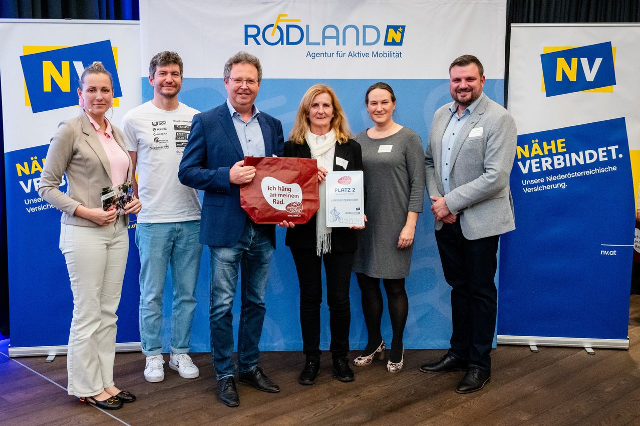 A group of people stand in front of a banner, two women hold awards, one woman holds a red bag, and a man holds a certificate.