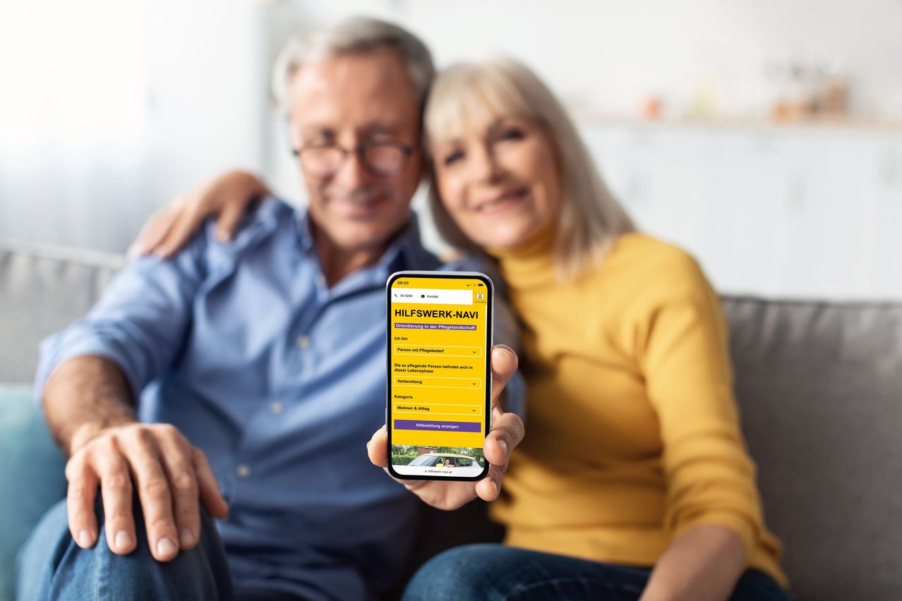 Ein älteres Paar sitzt auf einem Sofa, wobei der Mann ein Smartphone mit der App 'HILFSWERK-NAVI' in der Hand hält, die eine gelbe Benutzeroberfläche zeigt.
