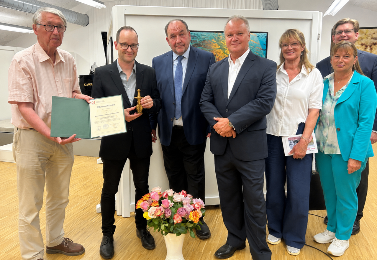 Fünf Personen stehen in einem Raum und posieren für ein Foto. Ein Mann hält ein Zertifikat und eine goldene Statuette. Die anderen drei Männer und eine Frau tragen Anzüge. Ein Blumenvase steht vor ihnen.