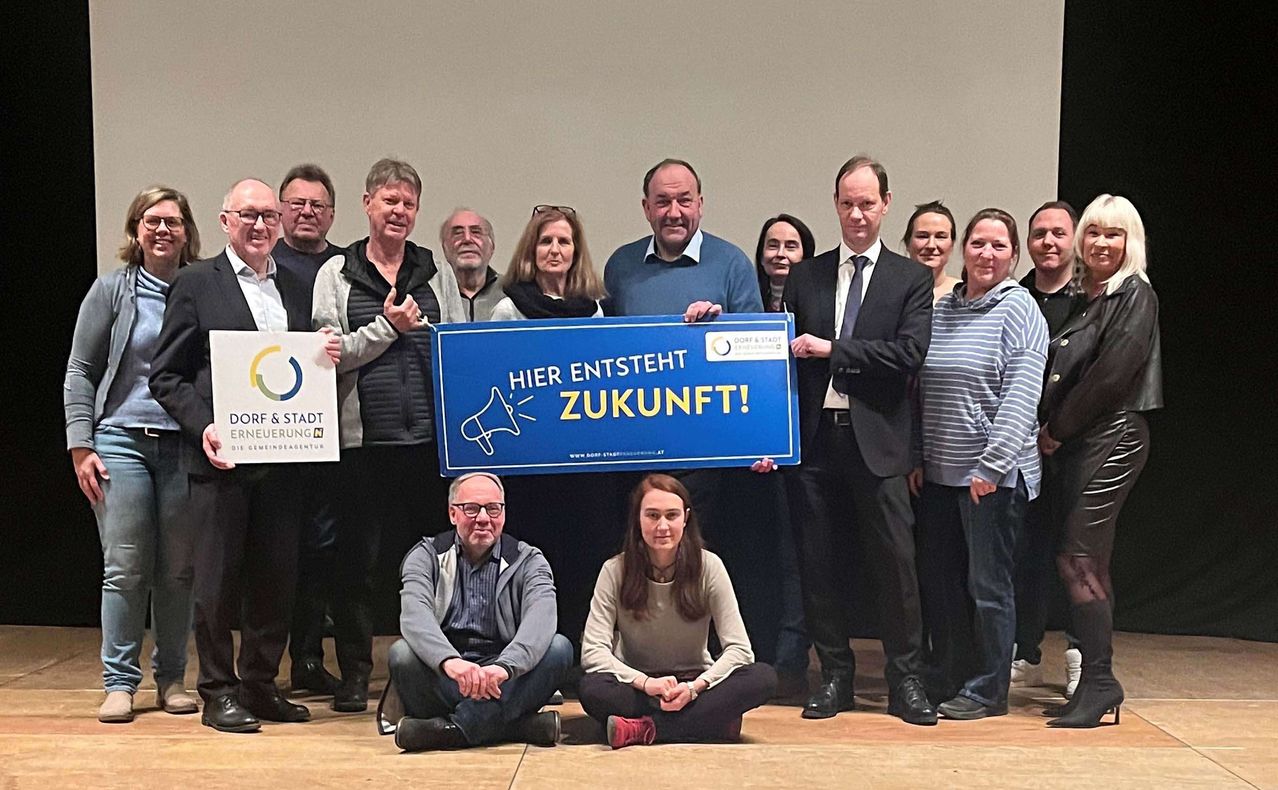 Eine Gruppe von Menschen steht und sitzt auf einer Bühne und hält ein Schild, auf dem 'Hier entsteht Zukunft!' steht. Zwei Personen halten das Schild hoch.