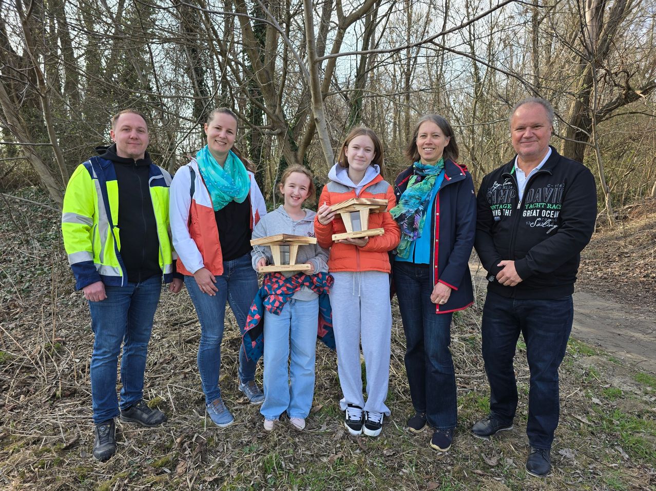 Fünf Personen stehen im Freien und lächeln. Zwei Mädchen halten Holzvogelhäuser, und andere tragen Jacken. Sie sind von kahlen Bäumen und Gras umgeben.