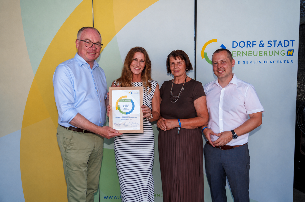 Four people are standing, smiling, and posing for a photo. A woman holds a certificate, and two men stand on either side. The woman wears a necklace, and the man on the right wears a wristwatch. The backdrop features the logo of 'DORNEUERNEUE'.
