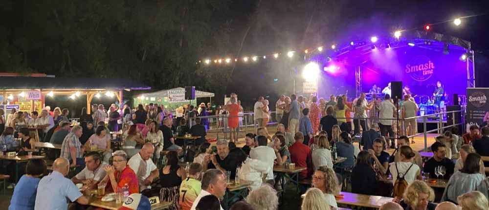 Eine Menschenmenge, die eine Veranstaltung im Freien bei Nacht genießt, mit aufgestellten Tischen und Stühlen, violetter und weißer Beleuchtung und einer Bühne mit einem Schild, auf dem Smash Time steht.