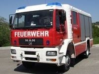 Ein roter und weißer Feuerwehrwagen steht am Straßenrand mit Bäumen im Hintergrund. Der Wagen hat das Wort 'FEUERWEHR' auf der Front.