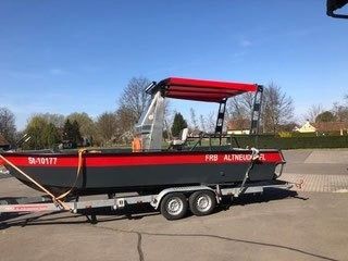 Ein rotes und schwarzes Boot mit der Nummer 9-10177 steht auf einem Anhänger auf einem Parkplatz. Das Boot hat ein rotes Verdeck und steht in einem Park mit Bäumen.