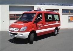 Ein roter und weißer Mercedes-Benz Sprinter Van, möglicherweise ein Feuerwehrfahrzeug, steht vor einem weißen Gebäude mit Glasfenstern und einer Garagentür.