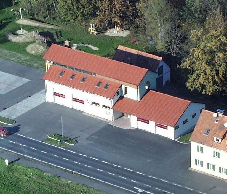 Ein Luftbild eines Gebäudes mit rotem Dach, mehreren Fenstern und einer Garage. Es ist von einem Rasen, Bäumen und einer Straße umgeben. Ein rotes Auto steht auf dem Parkplatz.