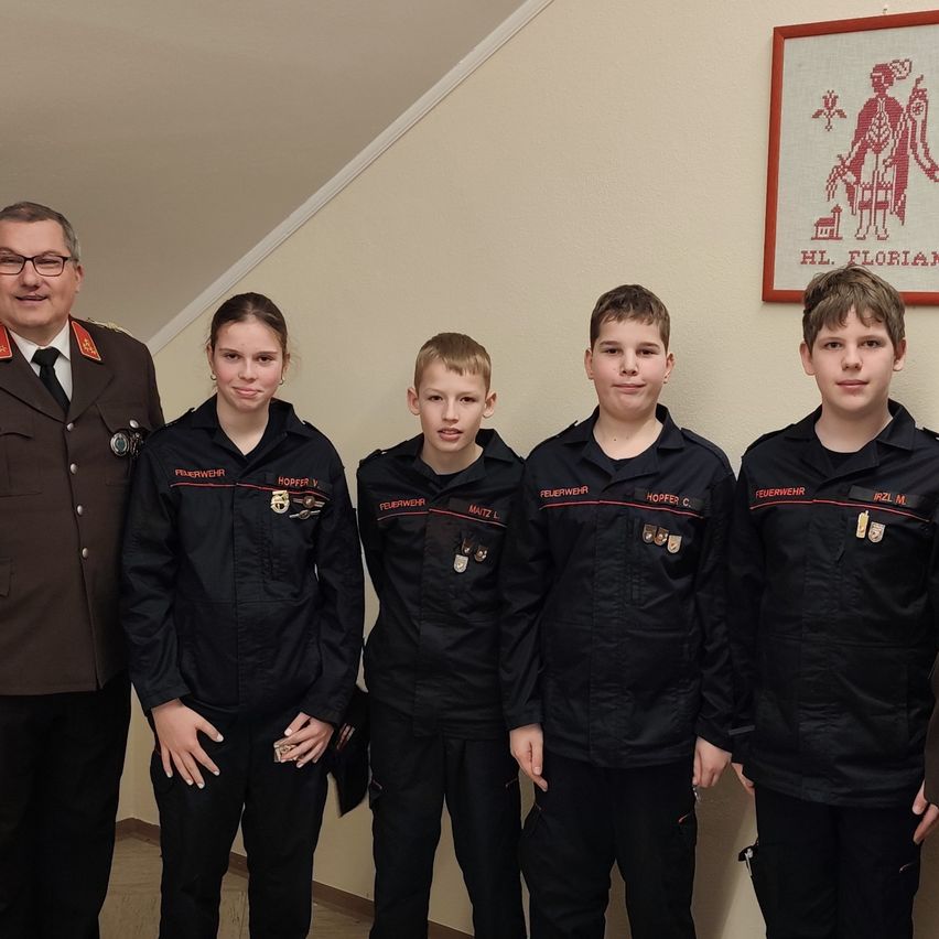 Fünf Personen in schwarzen Uniformen mit roten Abzeichen stehen zusammen. Ein Mann in Uniform steht hinter ihnen. Sie posieren für ein Foto vor einer Wand mit einem gerahmten Bild.