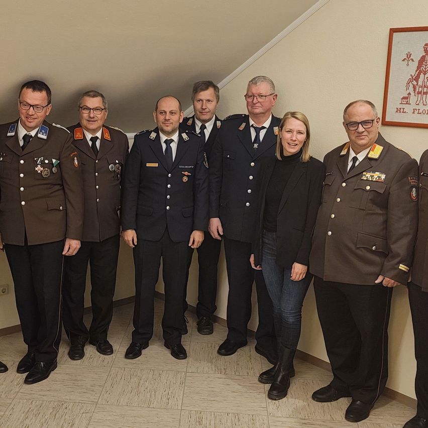 Sieben Personen in Militäruniformen stehen in einer Reihe und posieren für ein Foto. Darunter sind drei Männer und eine Frau mit Brille. Ein Bilderrahmen hängt an der Wand hinter ihnen.