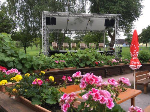 Ein Bühnenaufbau in einem Park mit Blumen, Lautsprechern und einem roten Schirm. Holzstühle sind auf der Bühne aufgestellt.