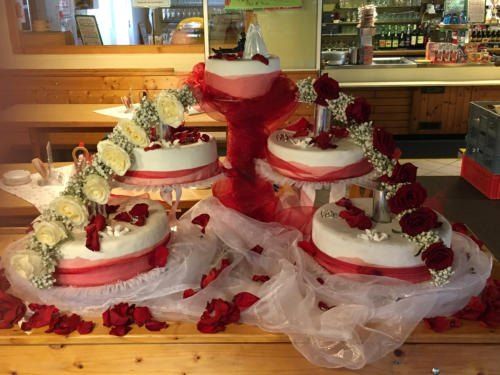 Vier Hochzeitstorten mit roten Bändern und Blumen auf einem Holztisch, umgeben von roten Blütenblättern.