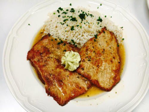 Ein Teller mit zwei gegrillten Fleischscheiben, einer Butterpatty und Reis garniert mit Petersilie.