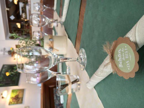 Ein Tischsetting mit Weingläsern, einem grünen Tuch und einem dekorativen Tag mit einem Tag, der Schon...DA BIST...? lautet. Der Tisch ist mit einem grünen Tuch bedeckt und hat ein weißes Tuch.