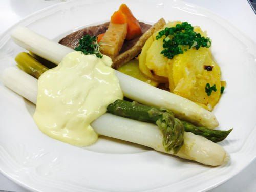 Ein weißer Teller mit einem Gericht aus weißen Spargel, Karotten, Kartoffeln und einer Sauce, garniert mit grünen Kräutern.