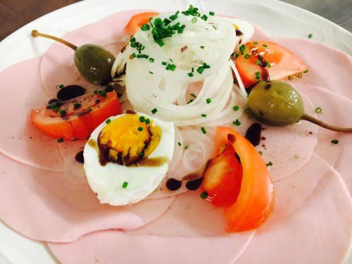 Ein Teller mit geschnittenem Schinken, halbiertem gekochtem Ei, Kirschtomaten, geschnittenen Zwiebeln und grünen Oliven, garniert mit Kräutern.