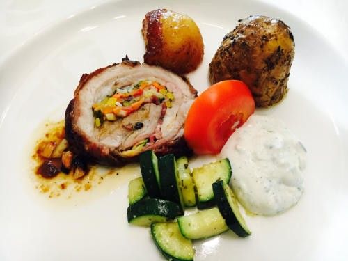 Ein weißer Teller mit einem Gericht, bestehend aus einem geschnittenen Schweinebraten gefüllt mit Gemüse, begleitet von gebratenen Kartoffeln, einer Tomate und einer weißen Sauce.