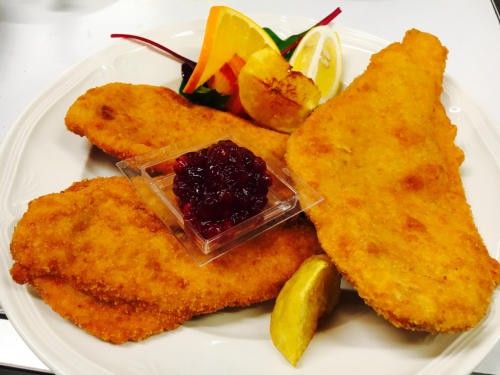 Ein weißer Teller präsentiert drei goldbraun gebratene Fischfilets, begleitet von einer kleinen Schüssel mit Cranberrysauce und Zitronenscheiben. Eine Garnierung aus Orangenscheiben und Kartoffelkegeln ist ebenfalls vorhanden.