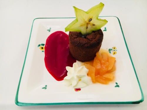 Ein quadratischer Teller zeigt einen Schokoladenmousse, gekrönt von Sternfrucht, roter Fruchtsauce, Schlagsahne und Orangenscheiben.