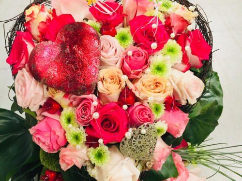 Ein herzförmiges Blumenarrangement mit roten Rosen, rosa Rosen und grünen Blättern. Das Herz ist mit rotem Glitzer und Perlen verziert.