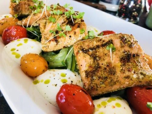 Ein weißer Teller mit gegrilltem Lachs, Kirschtomaten und Mozzarella-Kugeln. Das Gericht ist mit frischen Kräutern garniert.