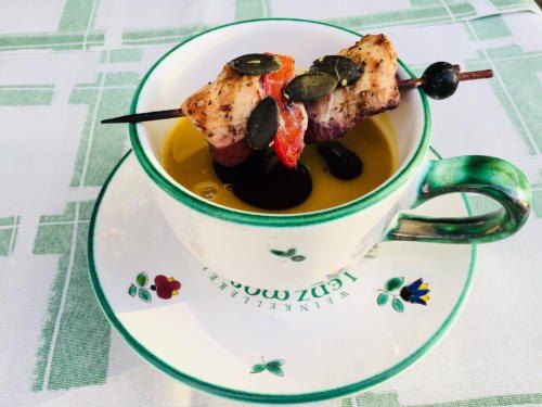 Eine Teetasse mit einer gelben Flüssigkeit enthält ein Spießstück Fleisch mit Tomaten und Oliven auf einer Untertasse mit Blumenmustern auf einem grün-weiß karierten Tischtuch.