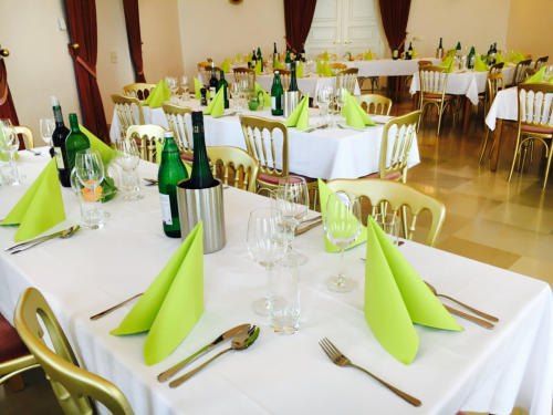 Ein Speisesaal ist für eine formelle Veranstaltung vorbereitet, mit Tischen, die mit weißen Tüchern bedeckt sind, geschmückt mit grünen Servietten, Weingläsern und Flaschen. Die Stühle sind golden mit Holzsitzen.