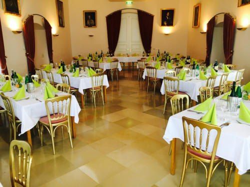 Ein Bankettsaal mit Tischen und Stühlen, die für eine formelle Veranstaltung vorbereitet sind. Jeder Tisch ist mit einer weißen Tischdecke bedeckt und hat ein Zentrum aus grünen Servietten. Der Raum hat einen gemusterten Boden und Wände, die mit Bilderrahmen geschmückt sind.