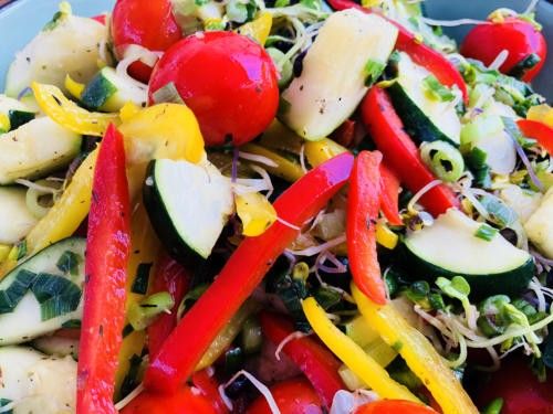 Ein farbenfroher Salat mit Kirschtomaten, Zucchini, gelben und roten Paprikaschoten und Kräutern, alles in einer Schüssel mit grünen Blättern angeordnet.