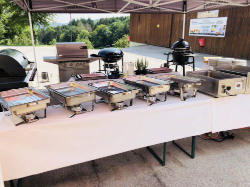 Ein Tisch unter einem Baldachin hat mehrere Speisewärmer und Grills. Die Speisewärmer sind aus Edelstahl mit Deckeln. Es gibt Teller, eine Topfpflanze und einen Feuerlöscher in der Nähe.