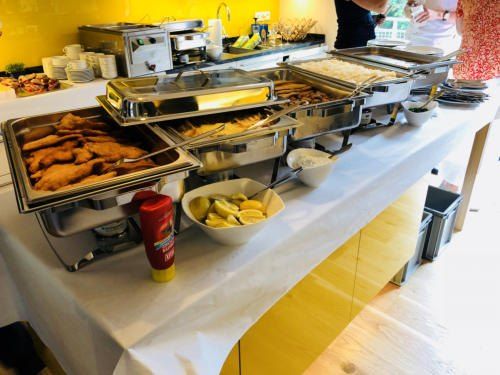 Ein Buffettisch mit Lebensmittelschalen, einer Schüssel mit Zitronen und einer Flasche Ketchup. Verschiedene Speisen werden in den Schalen serviert. Der Tisch ist mit einem weißen Tuch bedeckt.