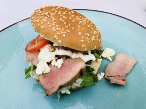 Ein Sandwich mit einem Sesambrötchen, belegt mit Scheiben rosa Fleisch, cremiger weißer Soße, Salat und Tomate, auf einem blauen Teller mit einem Stück Fleisch auf der Seite.