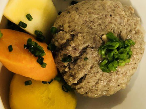 Ein Teller mit einem Fleischknopf, einer Karotte, einer Kartoffel und Frühlingszwiebeln. Der Fleischknopf hat eine braune Textur und grüne Zwiebeln sind darüber gestreut.