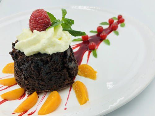 Ein Schokoladenkuchen mit weißer Creme, einer Himbeere und Minzblättern auf einem weißen Teller, garniert mit roter Beerensauce und Pfirsichscheiben.