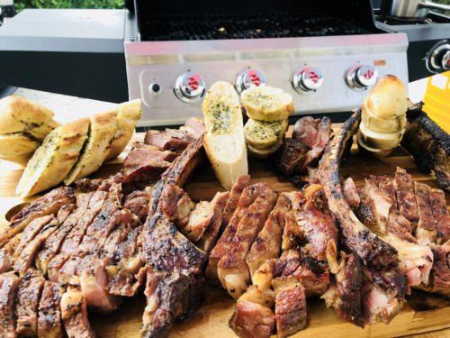 Gegrilltes Fleisch und Brot auf einem Schneidebrett vor einem Grill.