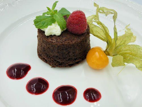 Ein Schokoladenkuchen auf einem weißen Teller, belegt mit Schlagsahne, einer Himbeere und Minzblättern. Fünf rote Saucenflecken und eine gelbe Frucht sind ebenfalls auf dem Teller.