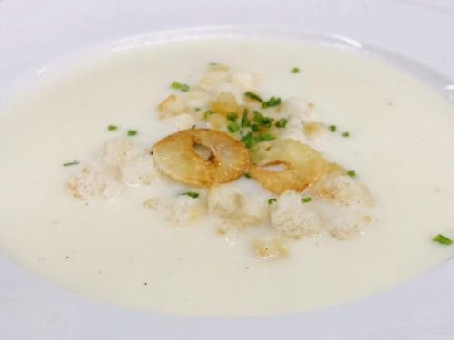 Eine Schüssel mit cremiger weißer Suppe, überzogen mit gerösteten Brotstücken und grünen Kräutern obenauf.