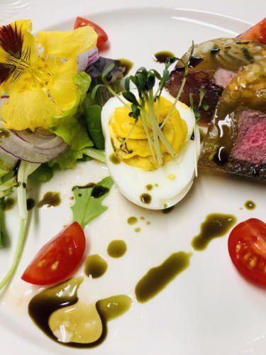 Auf einem weißen Teller befindet sich ein Gericht mit einem Rindersteak, einem pochierten Ei und einem Salat mit Tomaten und Blumen.
