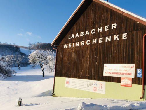 Ein holzverkleidetes Gebäude mit einem Schild, auf dem Laabacher Weinschenke steht, ist schneebedeckt. Im Hintergrund befindet sich ein Baum und ein Berg.