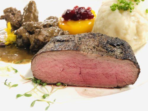 Ein Teller mit einem Steak und Kartoffelpüree. Das Steak ist angekohlt, und der Teller hat auch eine Preiselbeerensauce und eine Pfirsichscheibe.