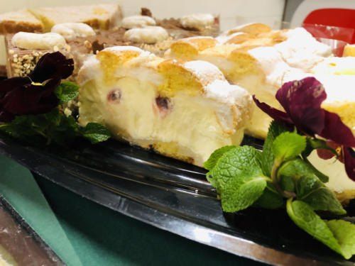 Eine Auswahl an Desserts ist auf einem schwarzen Tablett ausgestellt, darunter ein Stück Kuchen mit Sahne und Beeren, begleitet von Blumen und Minzblättern.