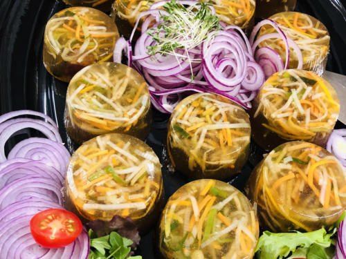 Ein schwarzer Teller zeigt geleeartige eingelegte Gerichte mit geriebenen Gemüsestücken, roten Zwiebeln und einer einzelnen roten Tomate, garniert mit Kräutern.