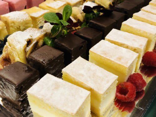 Eine Auswahl an Desserts wird ausgestellt, darunter geschichtete Kuchen mit weißer und brauner Glasur sowie andere, die mit Himbeeren und Minzblättern garniert sind.