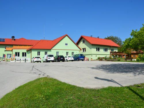 Ein grünes Gebäude mit roten Dächern und mehreren Fenstern. Ein Parkplatz mit mehreren geparkten Autos. Bäume und Pflanzen im Vordergrund.