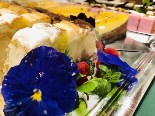 Eine Vielzahl von Desserts ist auf einem silbernen Tablett angeordnet, einschließlich einer blauen Blume, Himbeeren und Minzblättern. Sie sind auf einer reflektierenden Oberfläche ausgestellt.