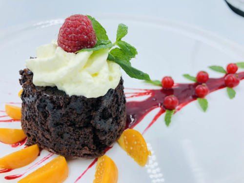 Ein Schokoladenkuchen, gekrönt mit Schlagsahne, einer Himbeere und Minzblättern. Er ist mit roter Sauce und Pfirsichscheiben garniert.