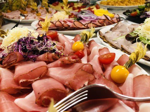 Eine Nahaufnahme eines Buffettisches, gefüllt mit verschiedenen Fleischsorten, Gemüse und Garnierungen. Eine Gabel ist im Vordergrund prominent zu sehen.