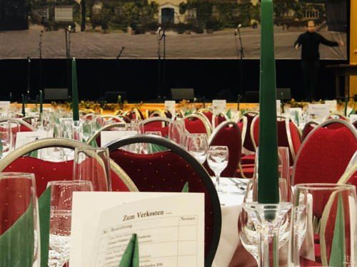 Ein Veranstaltungsraum mit großem Bühnenhintergrund und einem Künstler. Die Tische sind mit Weingläsern, Servietten und Menüs gedeckt. Der Raum ist mit roten Stühlen gefüllt.