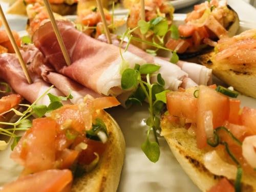 Eine Auswahl an Bruschettas mit Prosciutto-Scheiben, Tomaten und grünen Kräutern auf einem weißen Teller, garniert mit Zahnstochern.