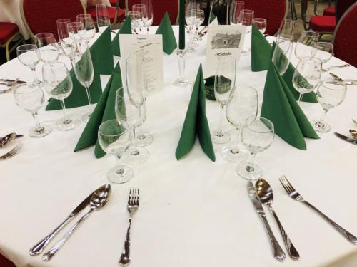 Ein gedeckter kreisförmiger Tisch für eine formelle Veranstaltung mit Weingläsern, Besteck, grünen Servietten und einer Speisekarte. Die Umgebung ist elegant mit weißen Tischdecken und roten Stühlen.